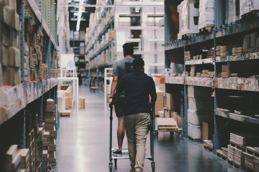 pexels-photo-1797428-1797428 Two men maneuver a trolley in a large warehouse filled with boxes and shelves.